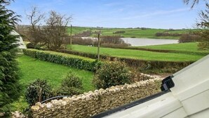 Une vue depuis une fenêtre donnant sur un paysage verdoyant, avec un mur de pierre et une étendue d’eau.
