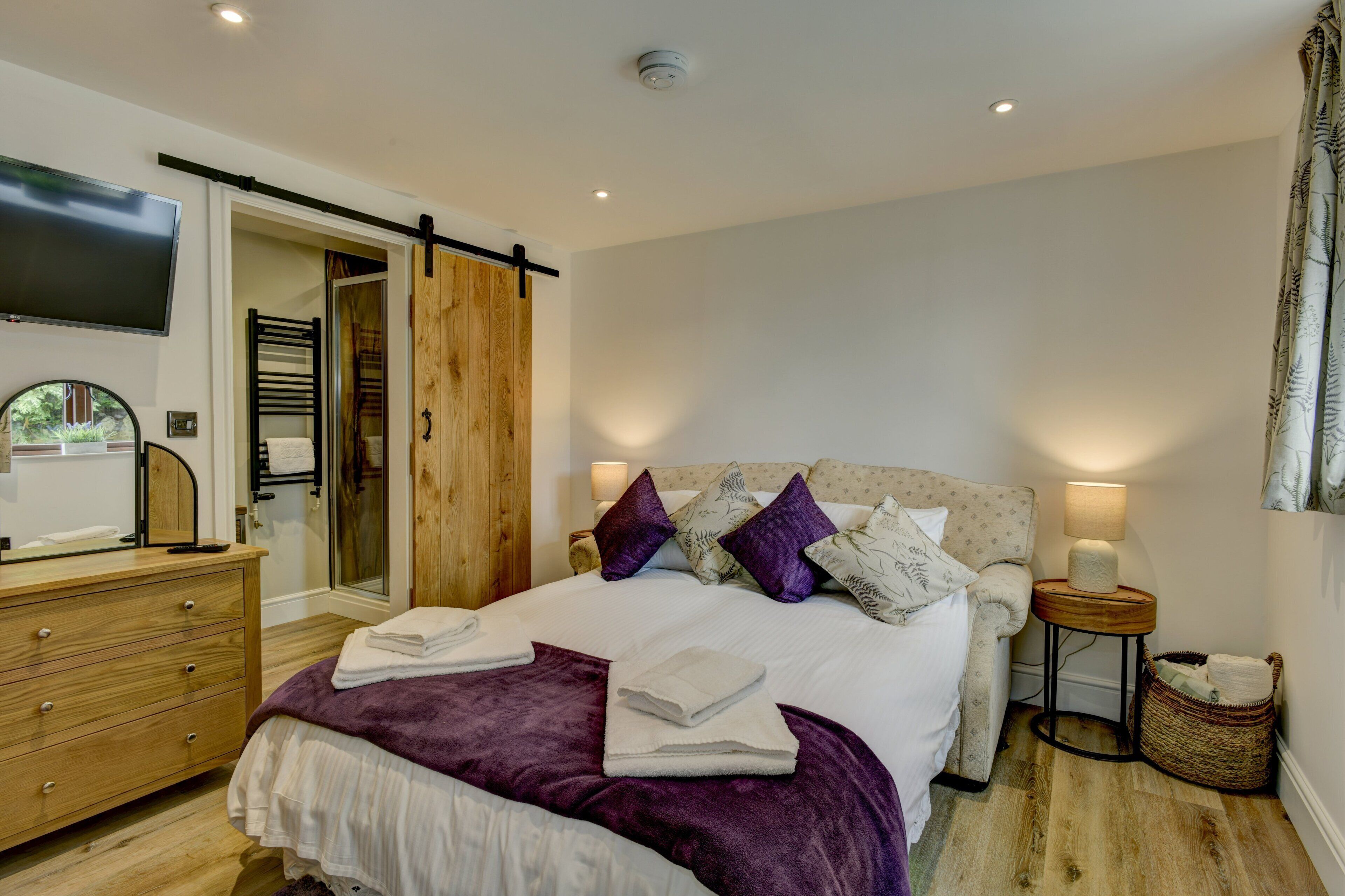 A bedroom with a bed, a dresser, a TV, a wooden door, and a small table with a lamp.