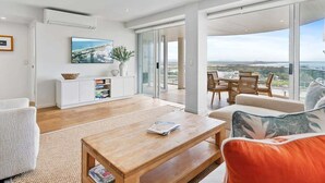 Amplia sala de estar con una mesa de centro de madera, un sofá con cojín, un televisor en la pared y un balcón con vistas al paisaje.