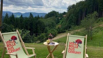Zwei Holzstühle mit aufgedruckter Schrift, ein kleiner Tisch mit Tasse und Blume, und eine schöne Aussicht auf einen bewaldeten Hügel.