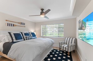 A bedroom with a bed, a ceiling fan, a window with blinds, a chair, and a large painting.