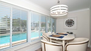 A dining area with a table and chairs, a clock on the wall, and a view of a pool through large windows. - RENOVATED 2 Bed, 2 Bath 1st floor with heated pool on the bay! (Naples)