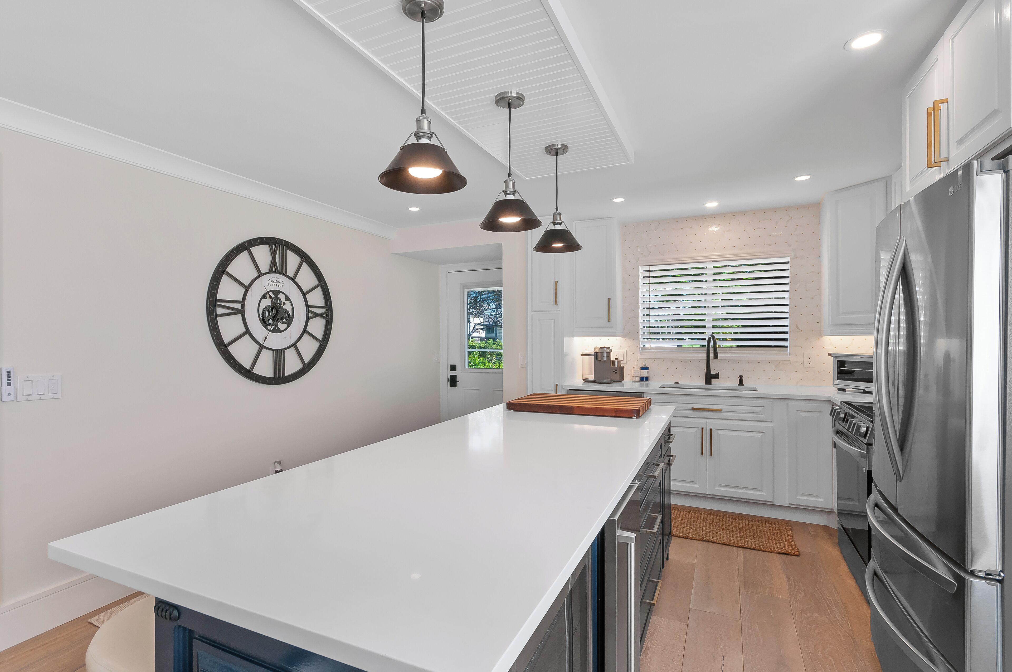 Una cocina moderna con armarios blancos, una isla grande, electrodomésticos de acero inoxidable, iluminación colgante y un reloj de pared.