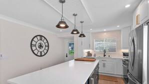 A modern kitchen with white cabinetry, a large island, stainless steel appliances, pendant lighting, and a wall clock. - RENOVATED 2 Bed, 2 Bath 1st floor with heated pool on the bay! (Naples)