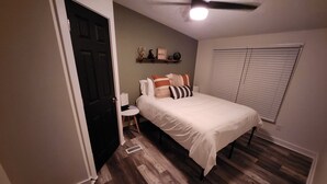 Une chambre à coucher comprenant un lit, une table de chevet, un ventilateur de plafond, une fenêtre avec des stores et une porte.