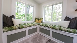 Un coin salon avec un banc, agrémenté d’un coussin en tissu à motifs, et flanqué de deux fenêtres donnant sur des arbres.
