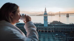 Una persona beu d'una tassa, amb una vista d'un edifici històric amb una torre del rellotge i un pont al fons.
