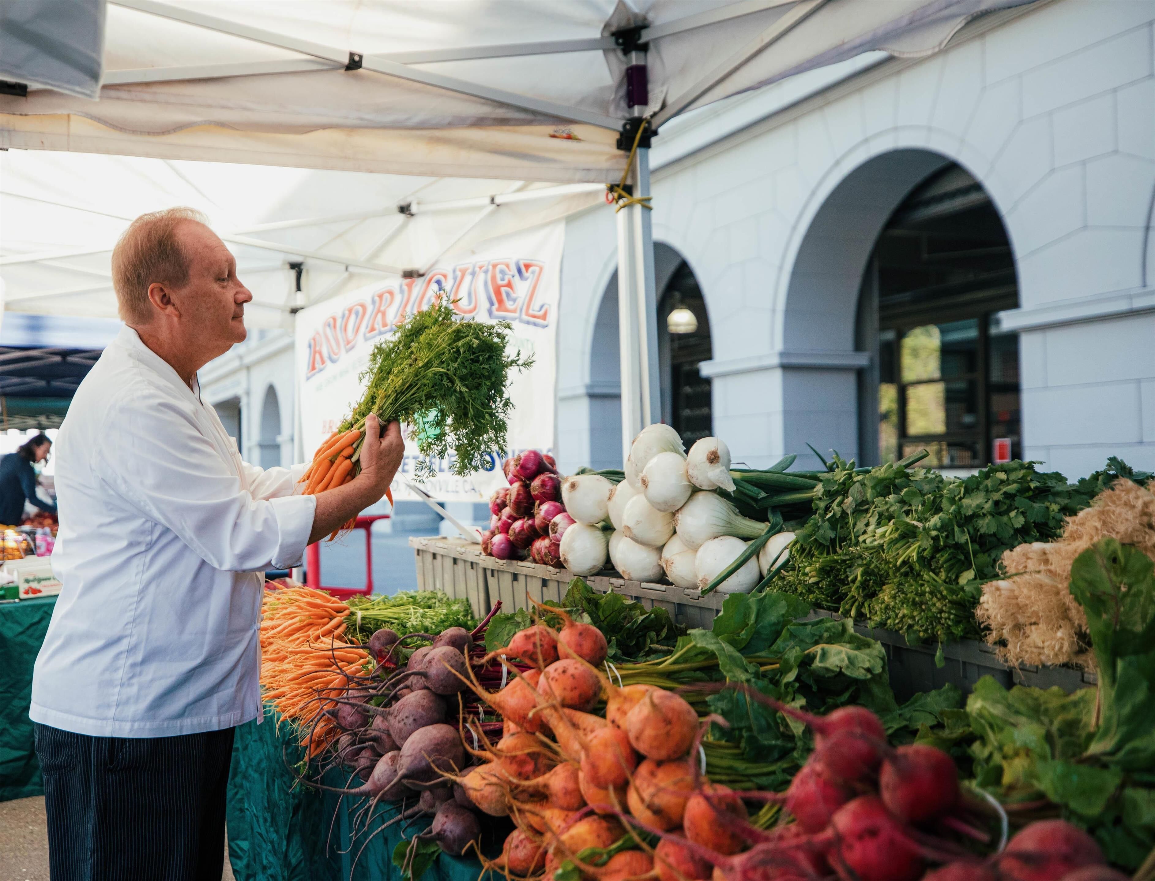 Un homme tenant un bouquet de carottes sur un étal de marché où sont exposés divers légumes.
