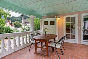 Une terrasse couverte avec une table et des chaises en bois, une porte blanche avec des panneaux de verre, et une vue sur une maison en arrière-plan.