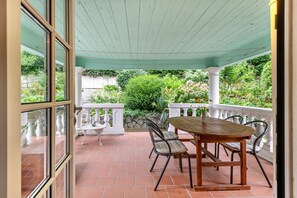 Une terrasse couverte avec une table et des chaises, des balustrades blanches et une vue sur la verdure.