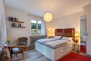 Une chambre avec un lit blanc, deux lampes de chevet, un tapis rouge, une chaise en osier et des étagères à livres.