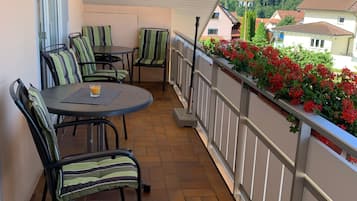 En balkon med et bord, stole, gelænder og udsigt til huse og grønne områder.