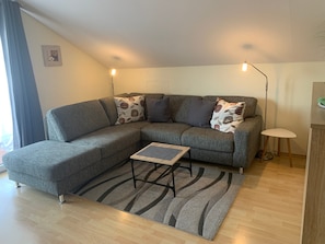 A modern living room with a grey sectional sofa, a small table, a patterned rug, and a floor lamp. - Apartment 'Haus Braun' with Shared Garden, Balcony, and Wi-Fi (Wasserburg)