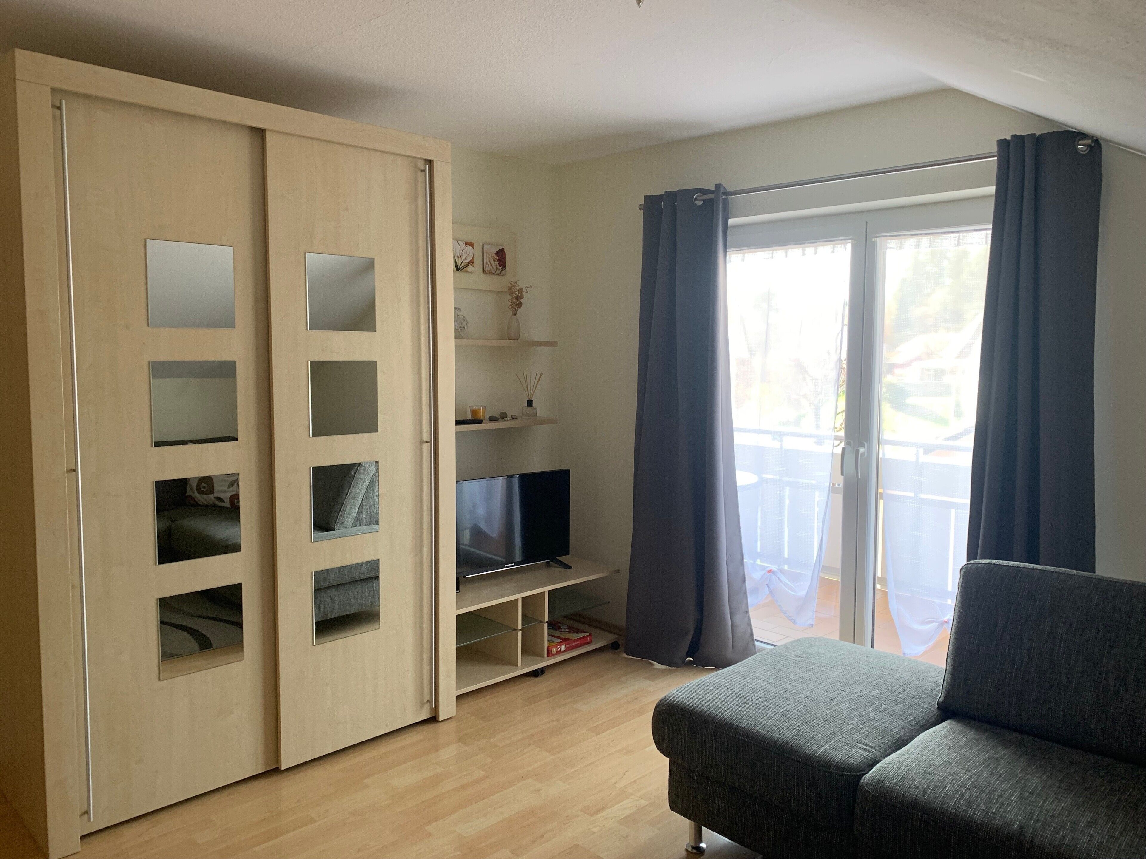 Ein Zimmer mit Schiebetürenschrank, Flachbildfernseher, grauem Sofa und Balkon mit Vorhängen.