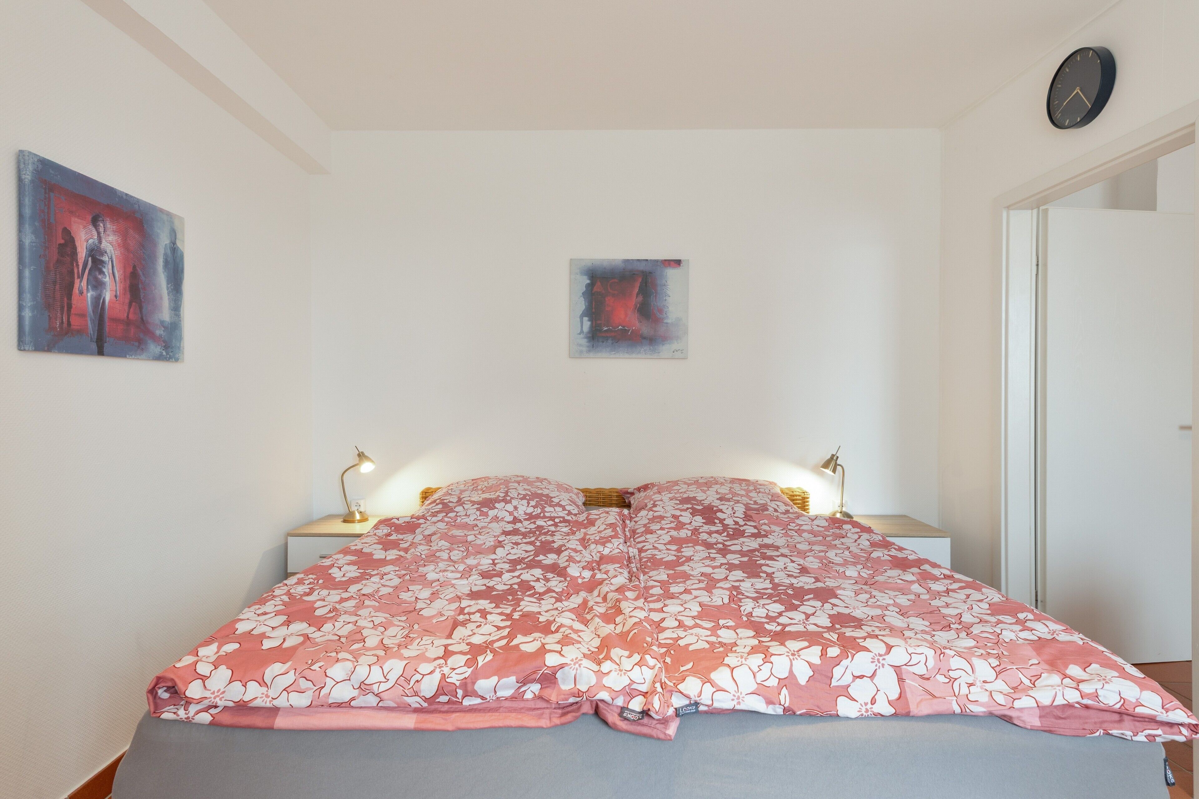 A bedroom with a bed, two bedside lamps, a wall clock, and two pieces of wall art.
