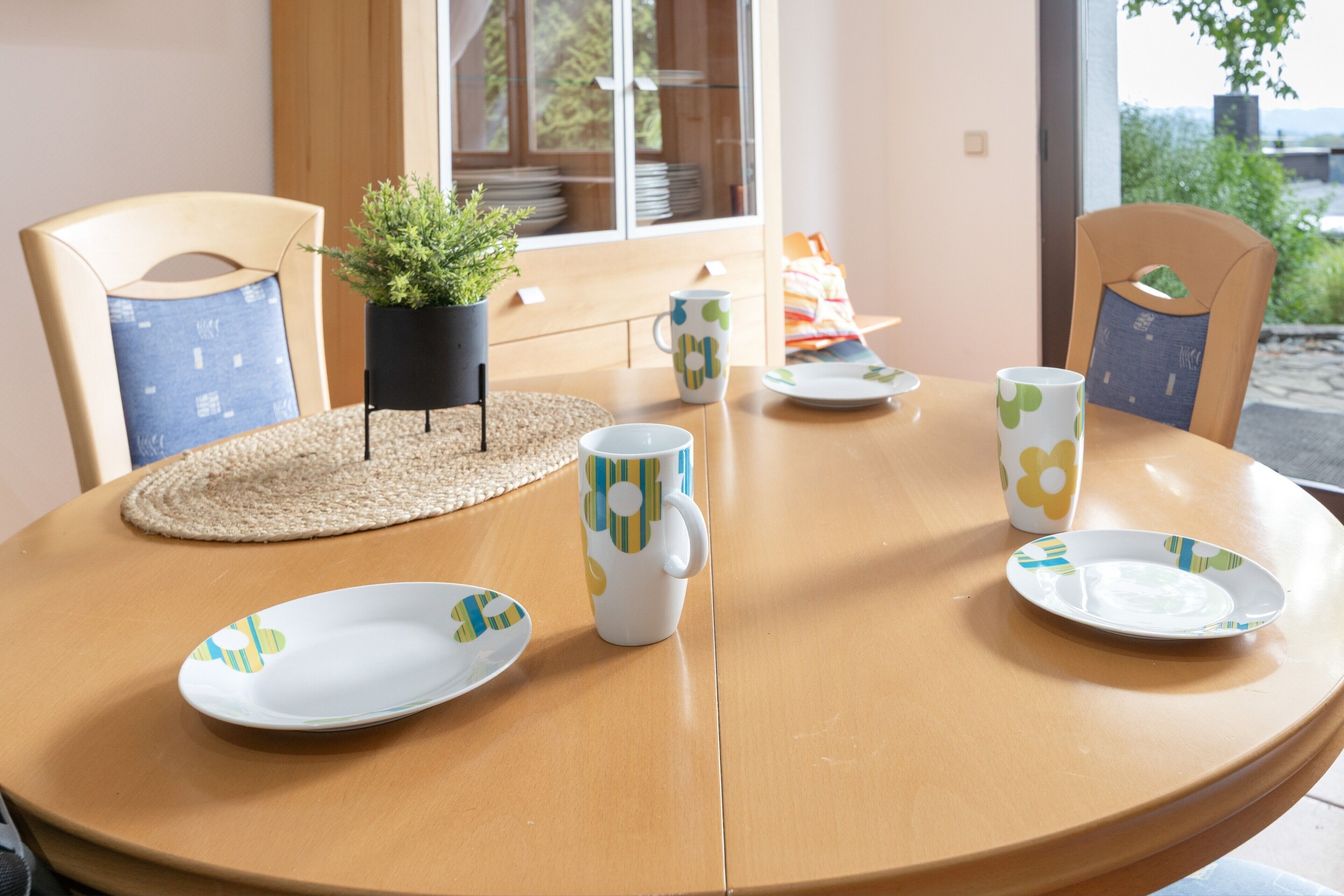 Une table ronde en bois, surmontée d’un set de table en fibres tressées, est entourée de chaises dotées de coussins bleus. Un pot de plante est placé sur le set de table.