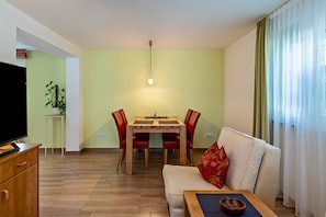Ein Wohnzimmer mit einer Couch, einem Esstisch mit Stühlen, einem Fernseher auf einer Holzkommode und einer Pflanze.