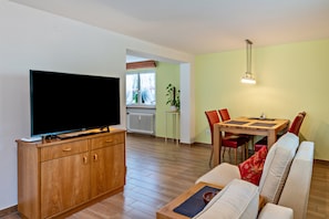 Ein Wohnzimmer mit Fernseher, Holzschrank, Sofa, Esstisch, Stühlen und einer Pendelleuchte.
