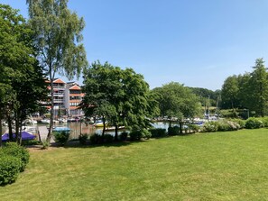 A grassy area with trees, a building with balconies, and boats in the background.