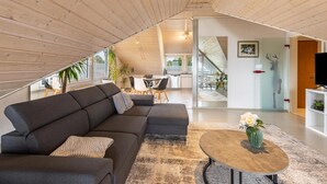 Ein modernes Loft mit Holzbalkendecke, einem dunkelgrauen Ecksofa, einem runden Couchtisch und einem Essbereich mit Glastür zum Küchenbereich.