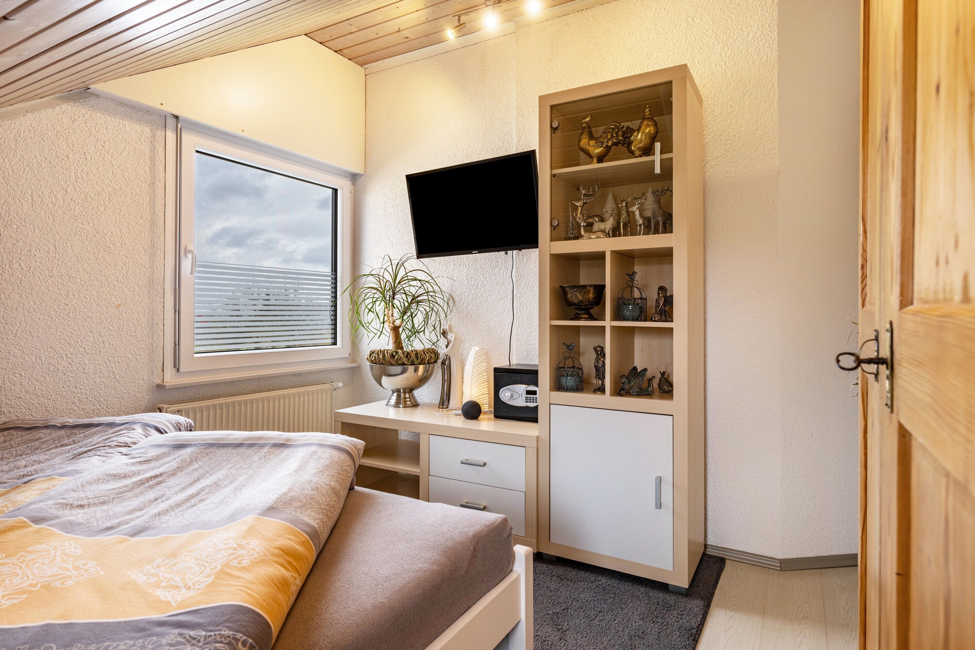 A bedroom with a bed, a desk with a computer, a TV, and a shelf with decorative items.