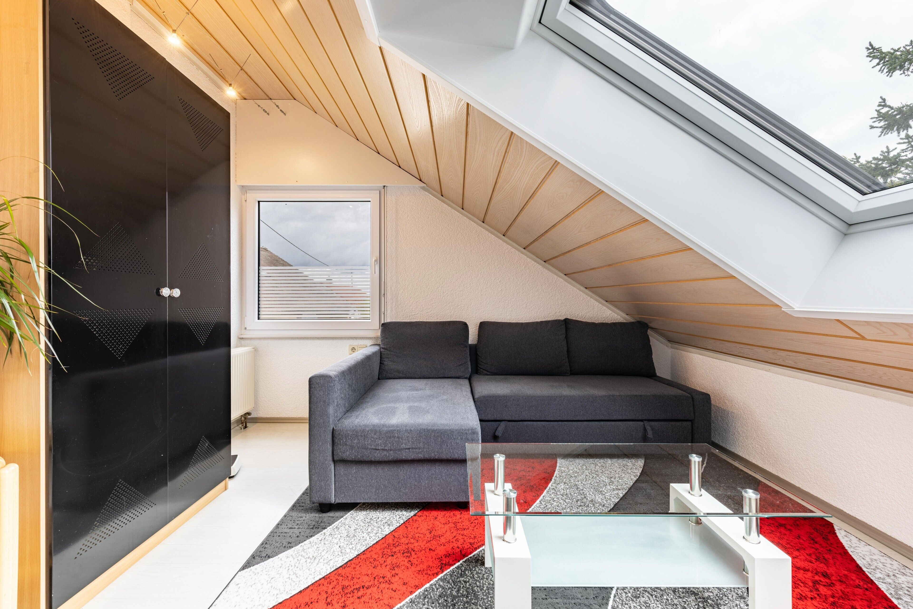 Une chambre de style loft moderne, avec un plafond en pente, une table basse en verre, un canapé d’angle gris et une grande fenêtre.