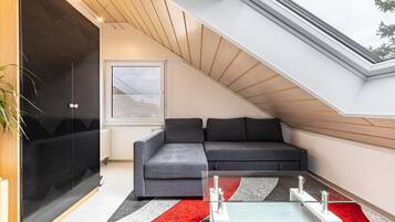 Une chambre de style loft moderne, avec un plafond en pente, une table basse en verre, un canapé d’angle gris et une grande fenêtre.