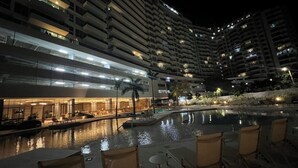 Ein Nachtblick auf ein Hotel mit Schwimmbad, Außenbereich zum Sitzen und mehreren Hochhäusern im Hintergrund.