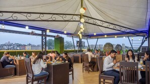 Een buitenrestaurant met tafels en stoelen onder een overdekte constructie, met stadsgezicht op de achtergrond.