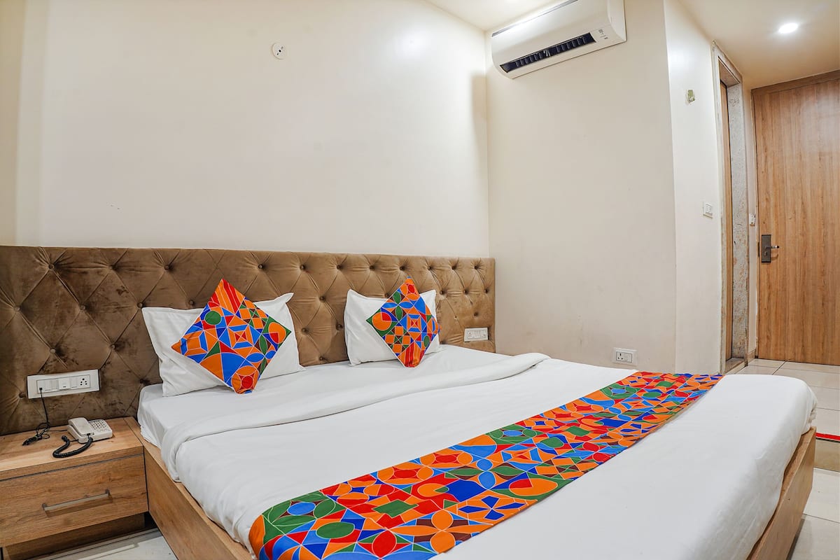 Una habitación de hotel con cabecera acolchada, cama con ropa blanca, dos almohadas coloridas, teléfono en la mesita de noche y una mesita de madera.