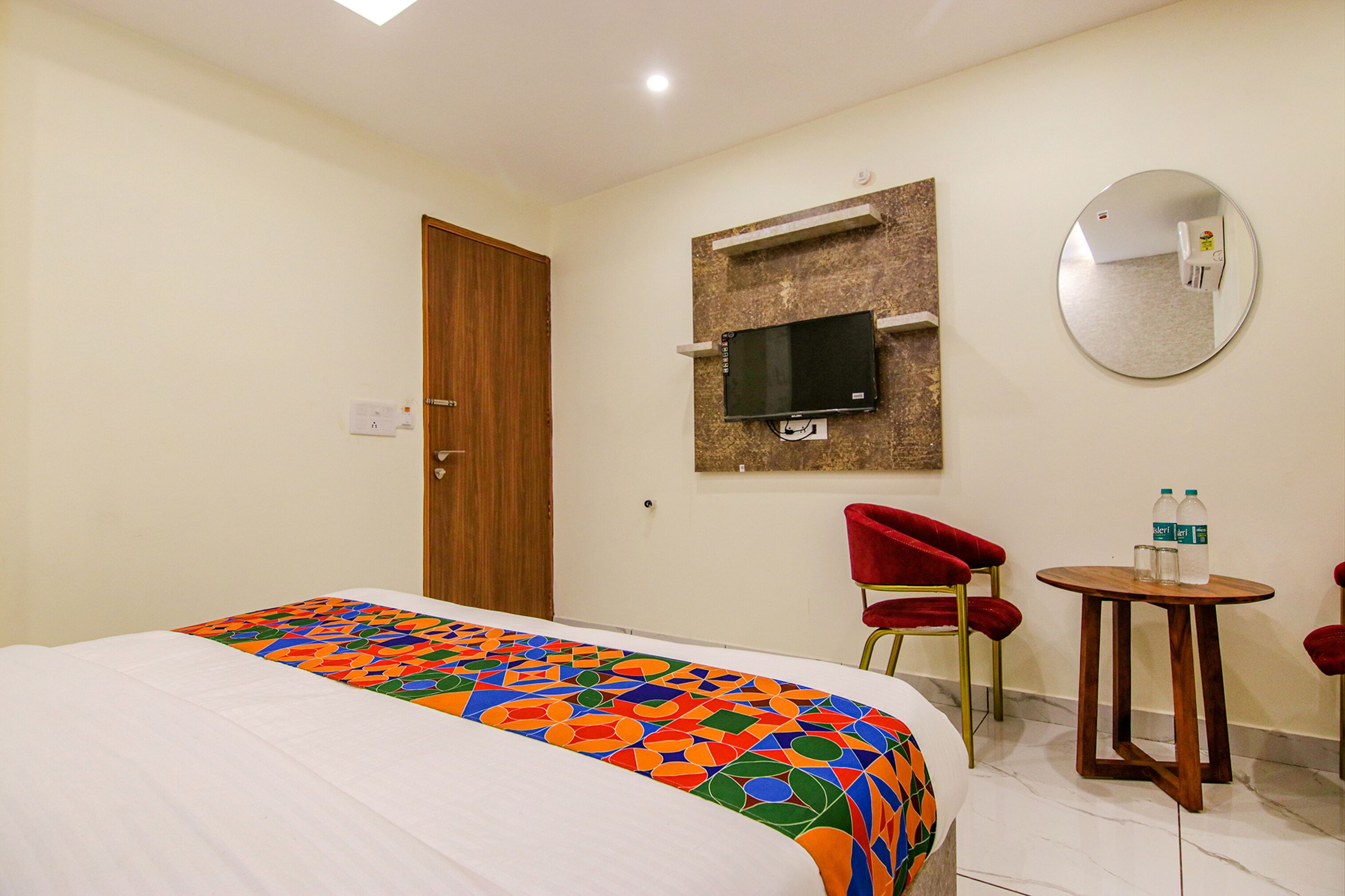 A hotel room with a bed, a TV mounted on the wall, a mirror, and two red chairs.