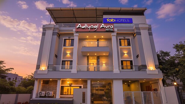 Um hotel moderno com telhado plano, várias varandas e uma placa chamativa para 'Adiyogi Aura Hotel'.