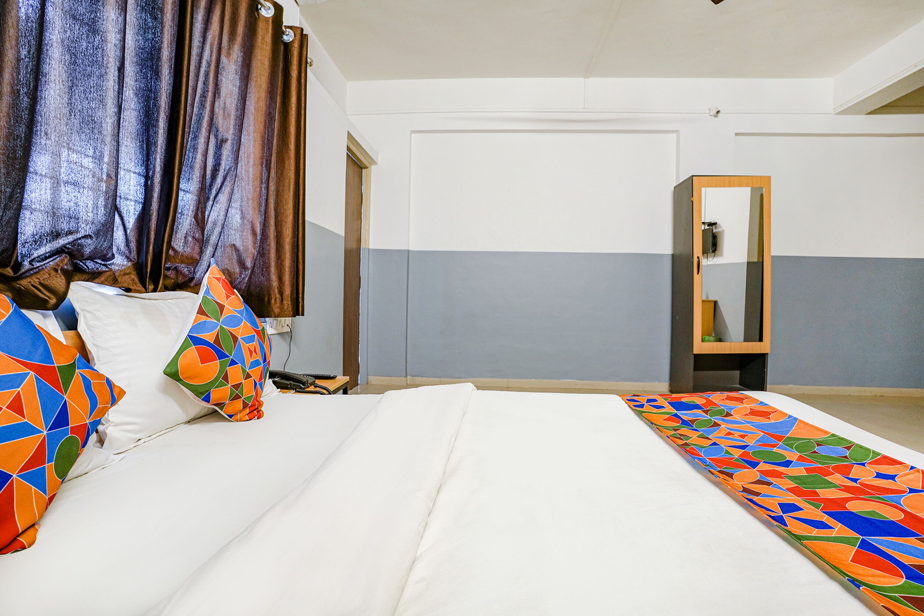 A hotel room with a bed, colorful pillows, a mirror, and a plain wall.