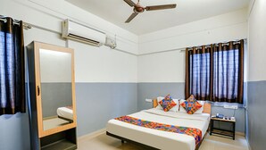 A bedroom with a bed, a bedside table, a mirror, a ceiling fan, and a window with curtains. - Fabhotel Bhakti Sankul (Nashik)