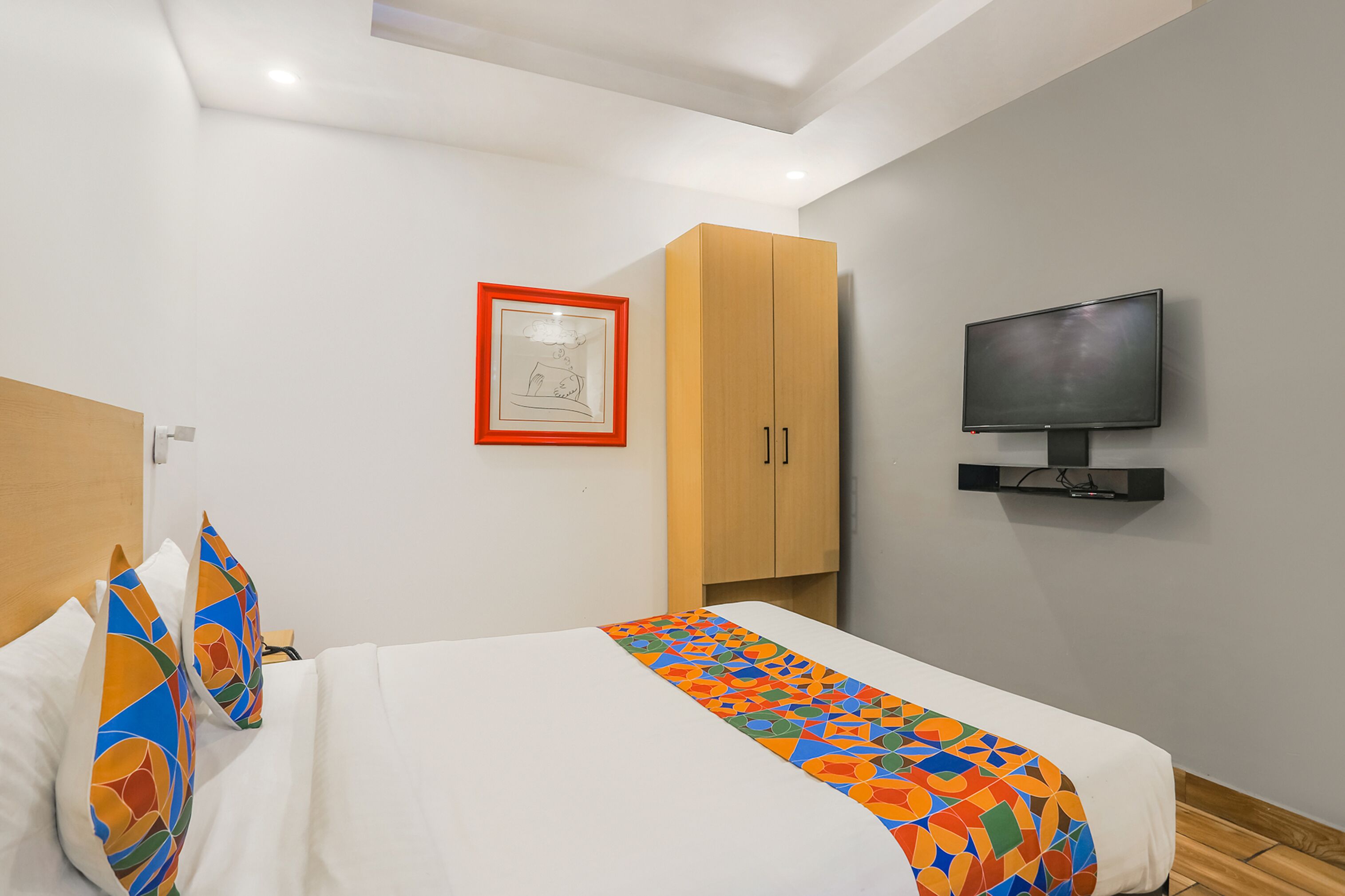 Une chambre d’hôtel avec un lit, un téléviseur fixé au mur, une armoire en bois et un tableau encadré accroché au mur.