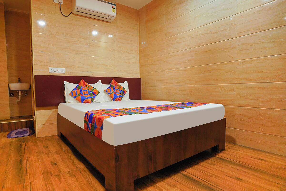 Una habitación de hotel con piso de madera, una cama con colchón blanco y almohadas de colores, cabecera de madera y aire acondicionado instalado en la pared.