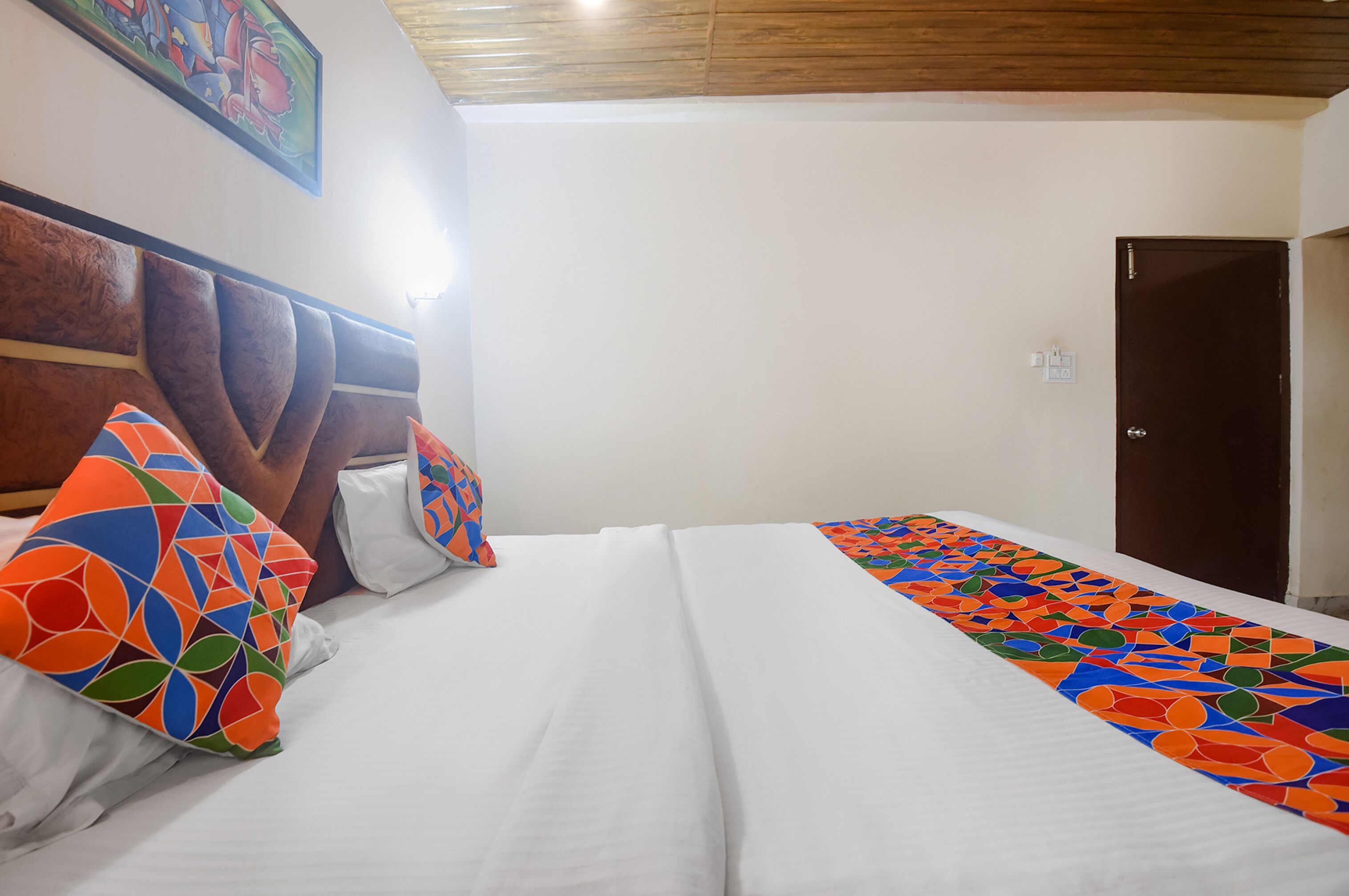 A hotel room with a bed, pillows, a headboard, a door, and a wall with a painting.