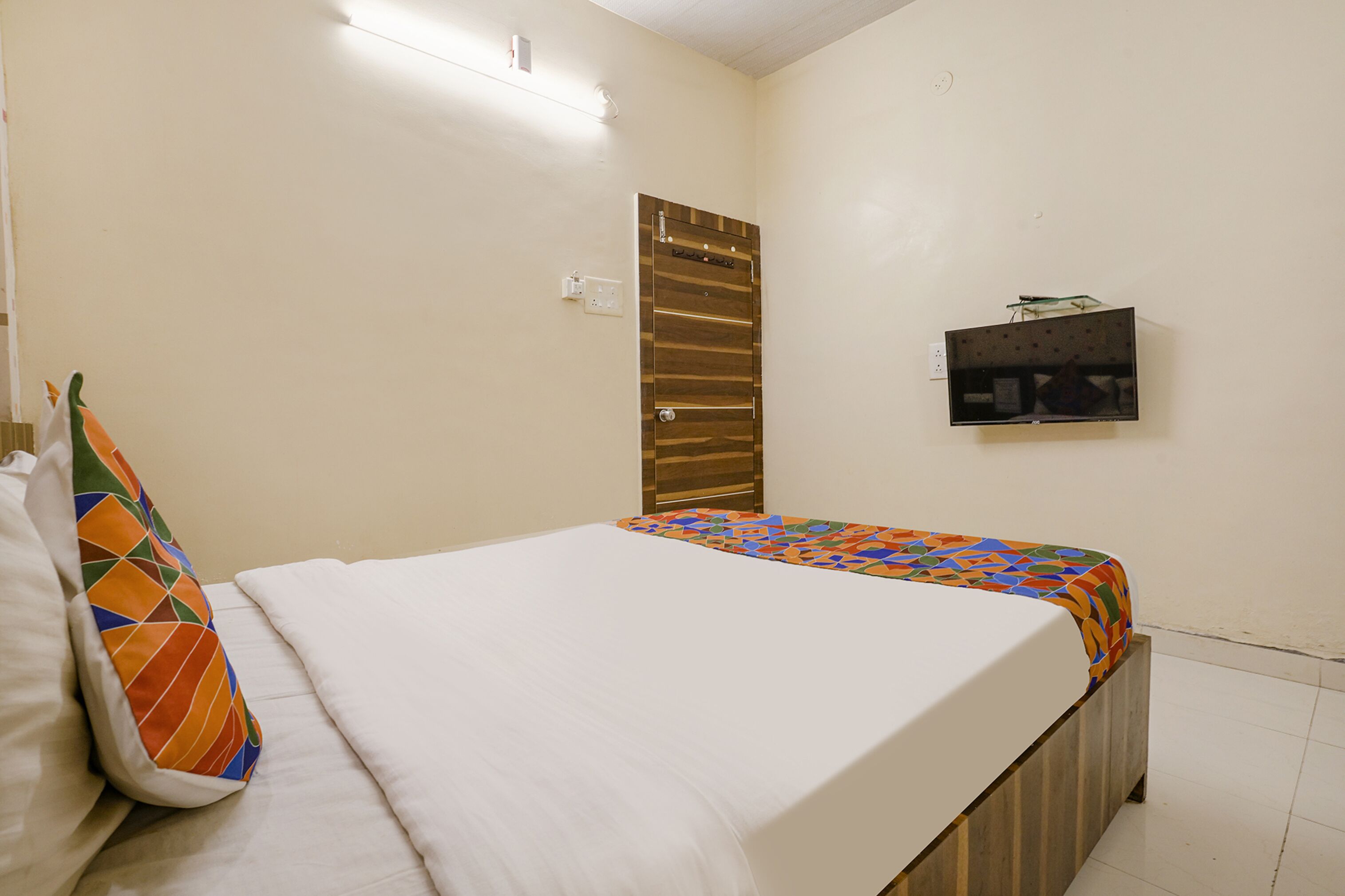 Una cama bien hecha con un edredón colorido y una sola almohada, una puerta de madera y un estante pequeño con objetos en la pared.