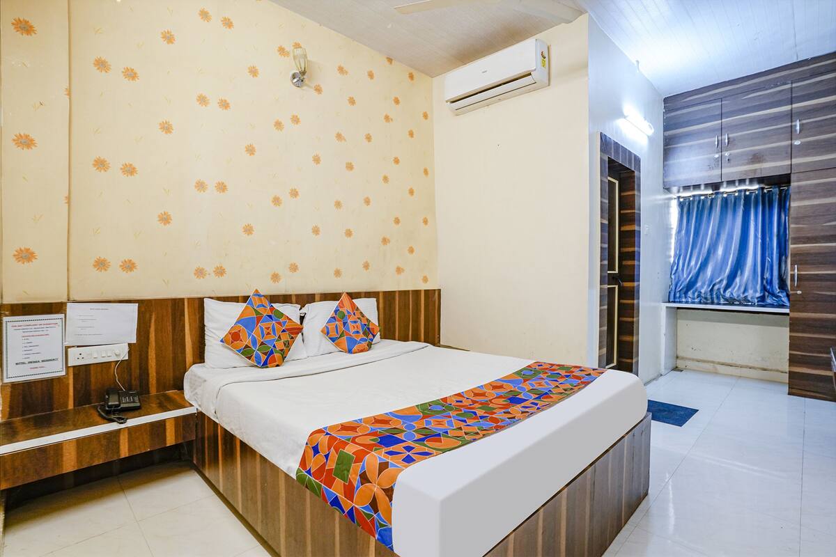 A hotel room with a wooden headboard, a bed with a colorful geometric bedspread, a floral-patterned wall, and an air conditioning unit.