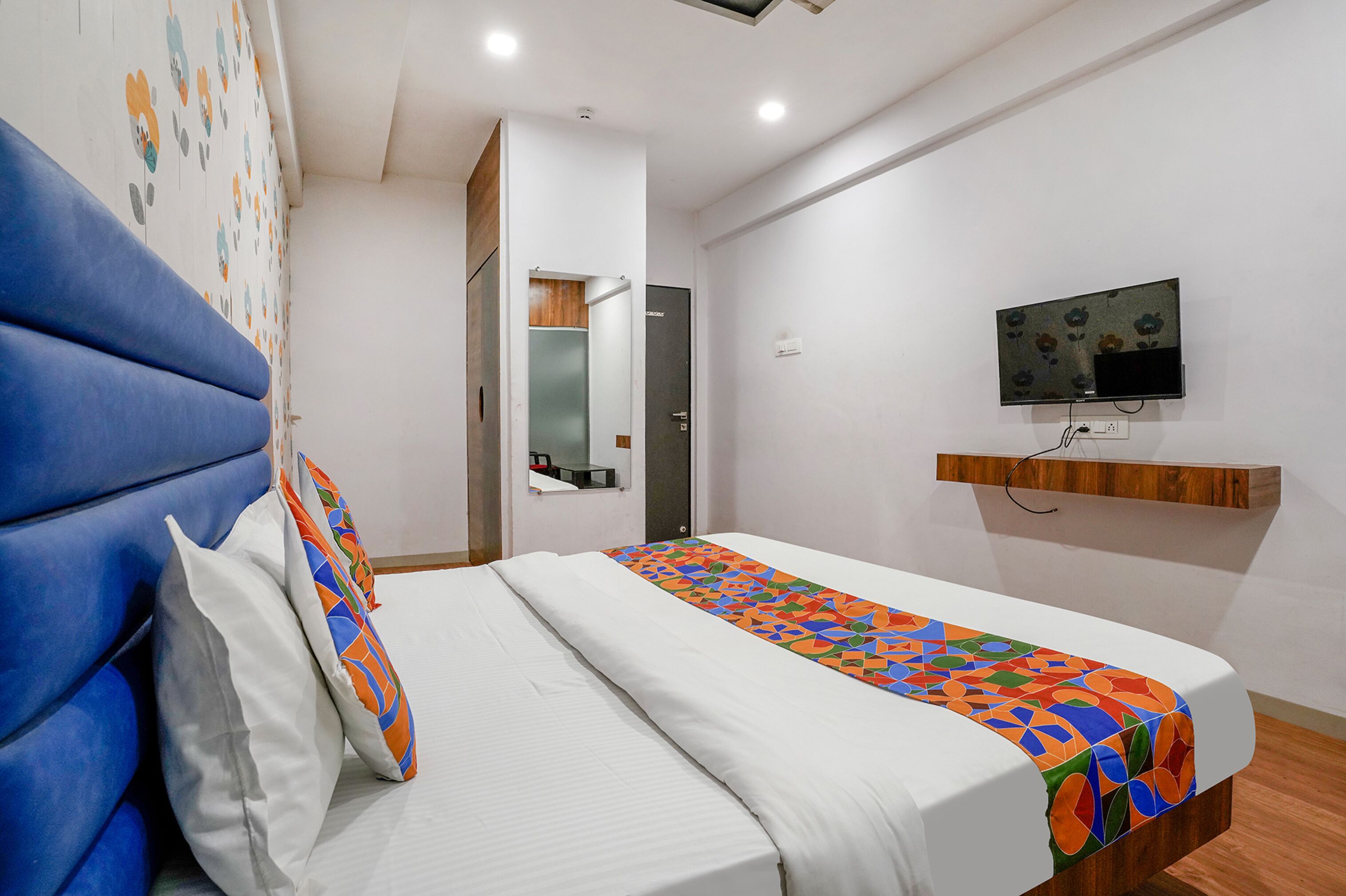 Una camera d'hotel con un letto, una testiera blu, una TV a muro e una mensola in legno.