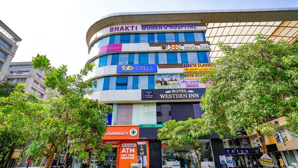 Un edificio de varios pisos con HOSPITAL BHAKTI WOMEN’S en la parte superior, servicios de atención dental y diversas tiendas, incluyendo una farmacia y un hotel llamado Western Inn.