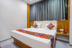 A neatly made bed with a colorful bedspread, a wooden headboard, and a bedside table with a red bottle. - Fabhotel Western Inn (Surat)
