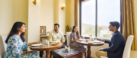 Un comedor con cuatro personas sentadas alrededor de una mesa, una ventana con cortinas, y una lámpara fijada a la pared.