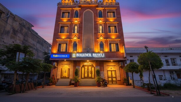 Un edificio con un letrero que indica 'Bikaner Haveli' y 'FabHotels'. Tiene un gran arco central, varias ventanas y está rodeado de árboles y vehículos estacionados.