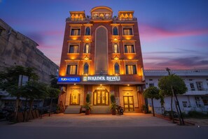 Un edificio con un letrero que indica 'Bikaner Haveli' y 'FabHotels'. Tiene un gran arco central, varias ventanas y está rodeado de árboles y vehículos estacionados.