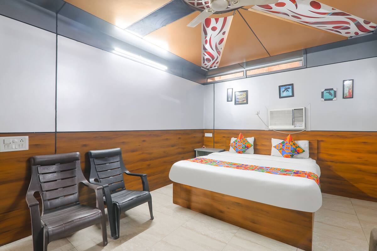 Quarto de hotel com cabeceira de madeira, cama com colcha colorida, duas cadeiras, ventilador de teto e ar condicionado fixado na parede.