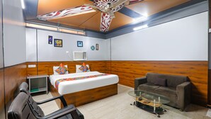 A hotel room with a bed, a sofa, a glass coffee table, a desk with a chair, and a ceiling fan. - Fabexpress Atithi (Nagri)