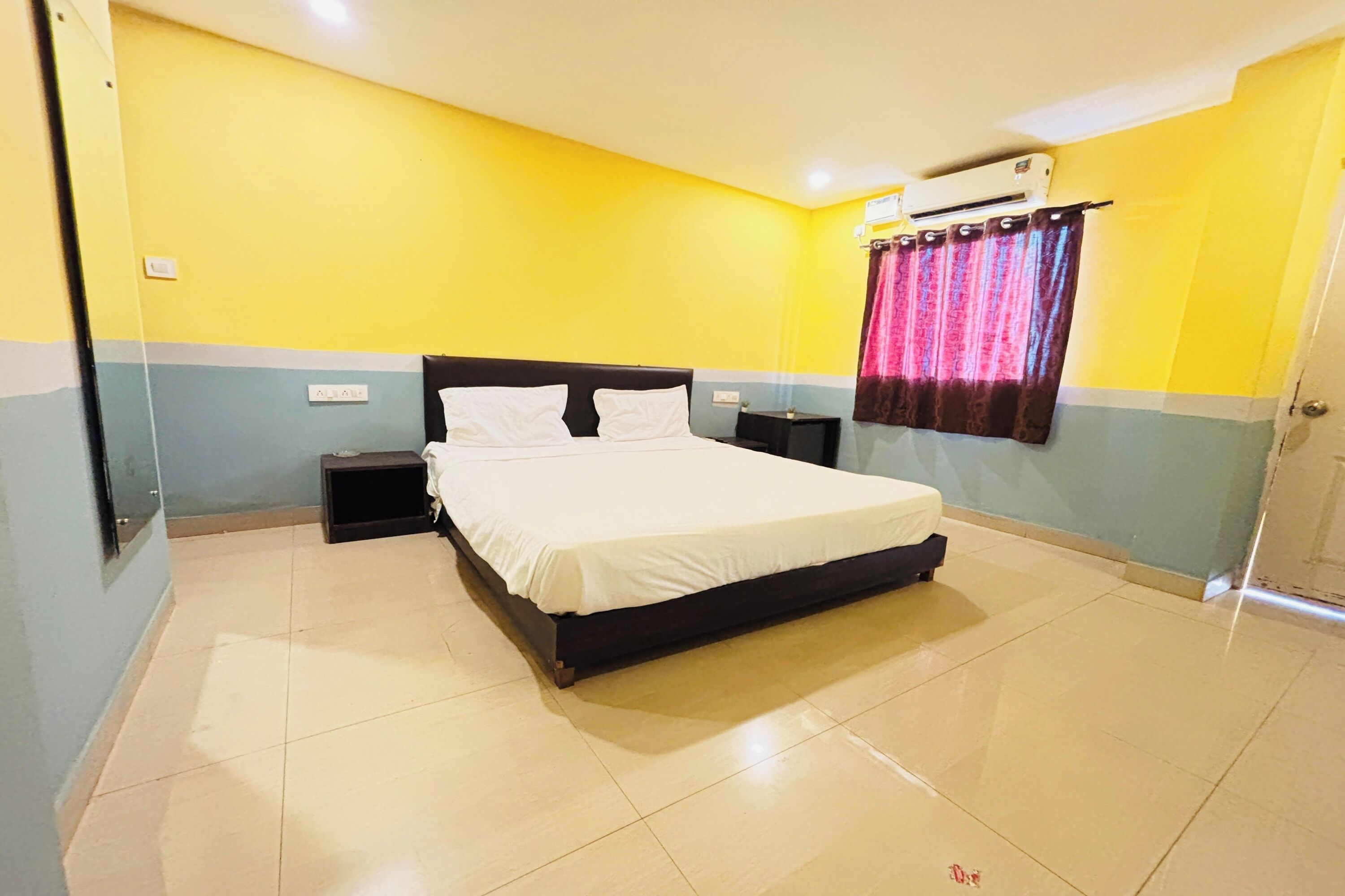 A hotel room with a bed, nightstands, an air conditioner, and a curtain.