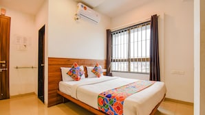 Habitación de hotel con una cama de madera, ropa de cama blanca, un edredón colorido, una ventana con cortinas y un equipo de aire acondicionado.