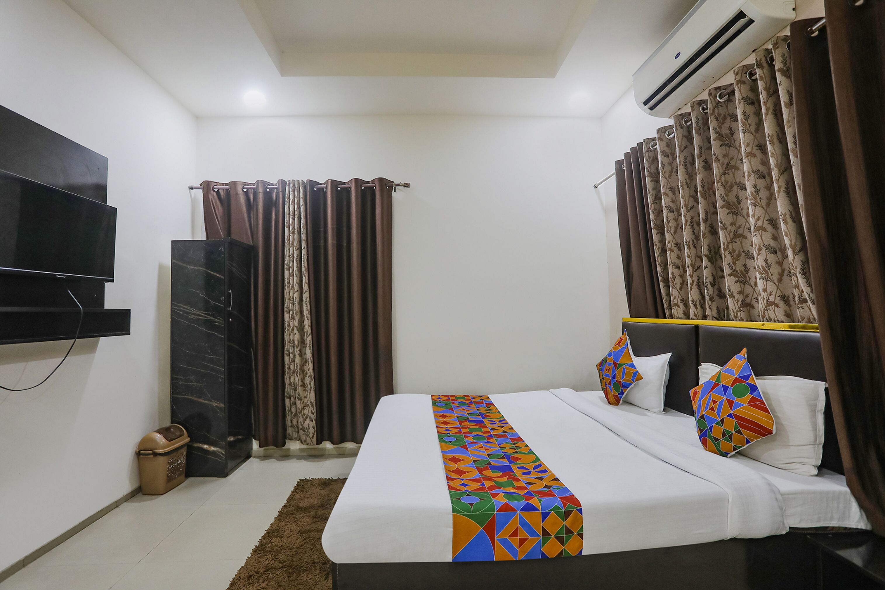 A hotel room with a bed, a flat-screen TV mounted on the wall, an air conditioning unit, and a window with curtains.